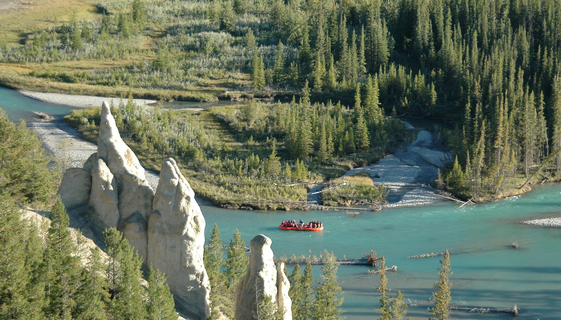 An orange raft with passengers floating the turquoise Bow River past hoodoo rock formations and evergreen forest
